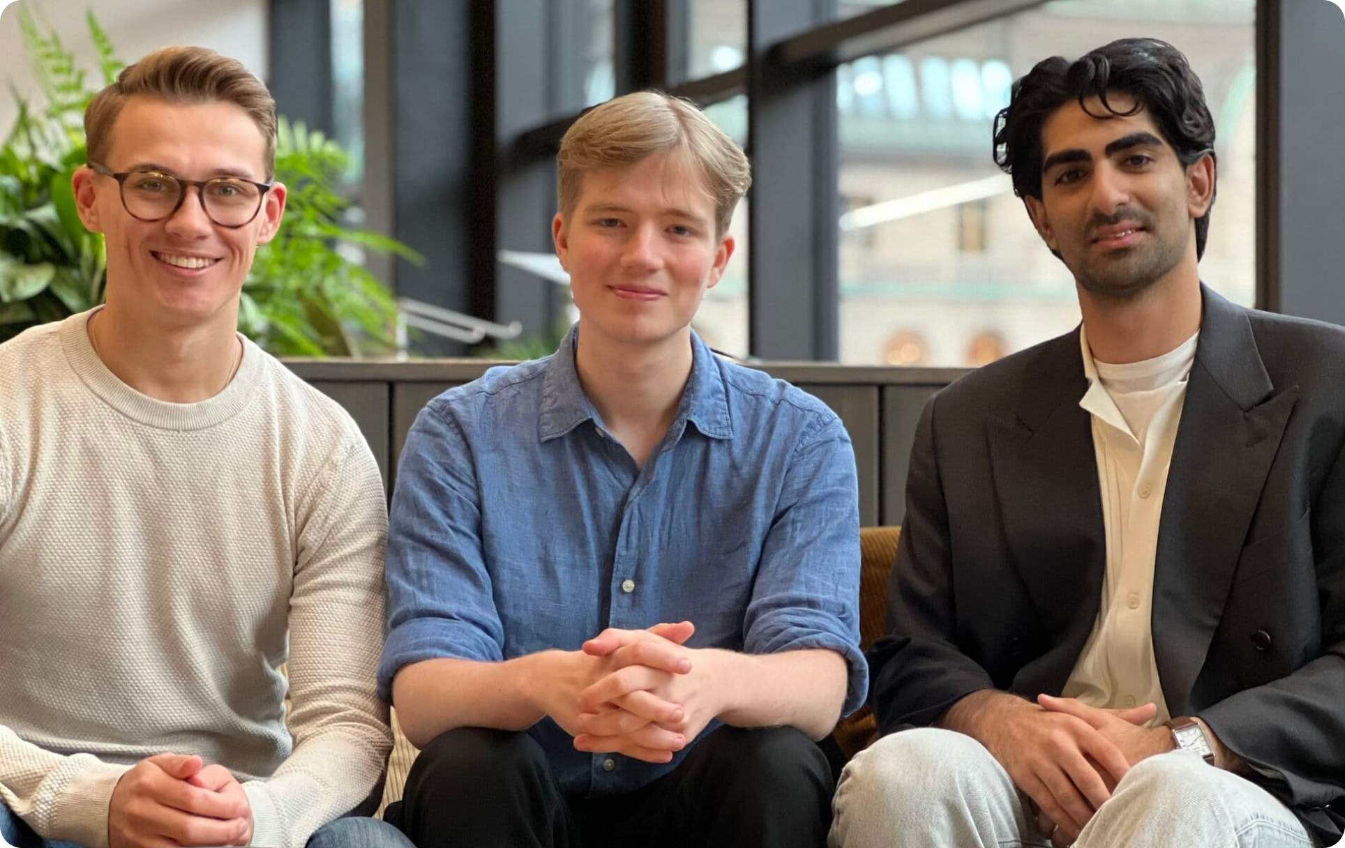 Three individuals sit closely together on a sofa, dressed in casual and smart attire, surrounded by greenery in a modern indoor space.